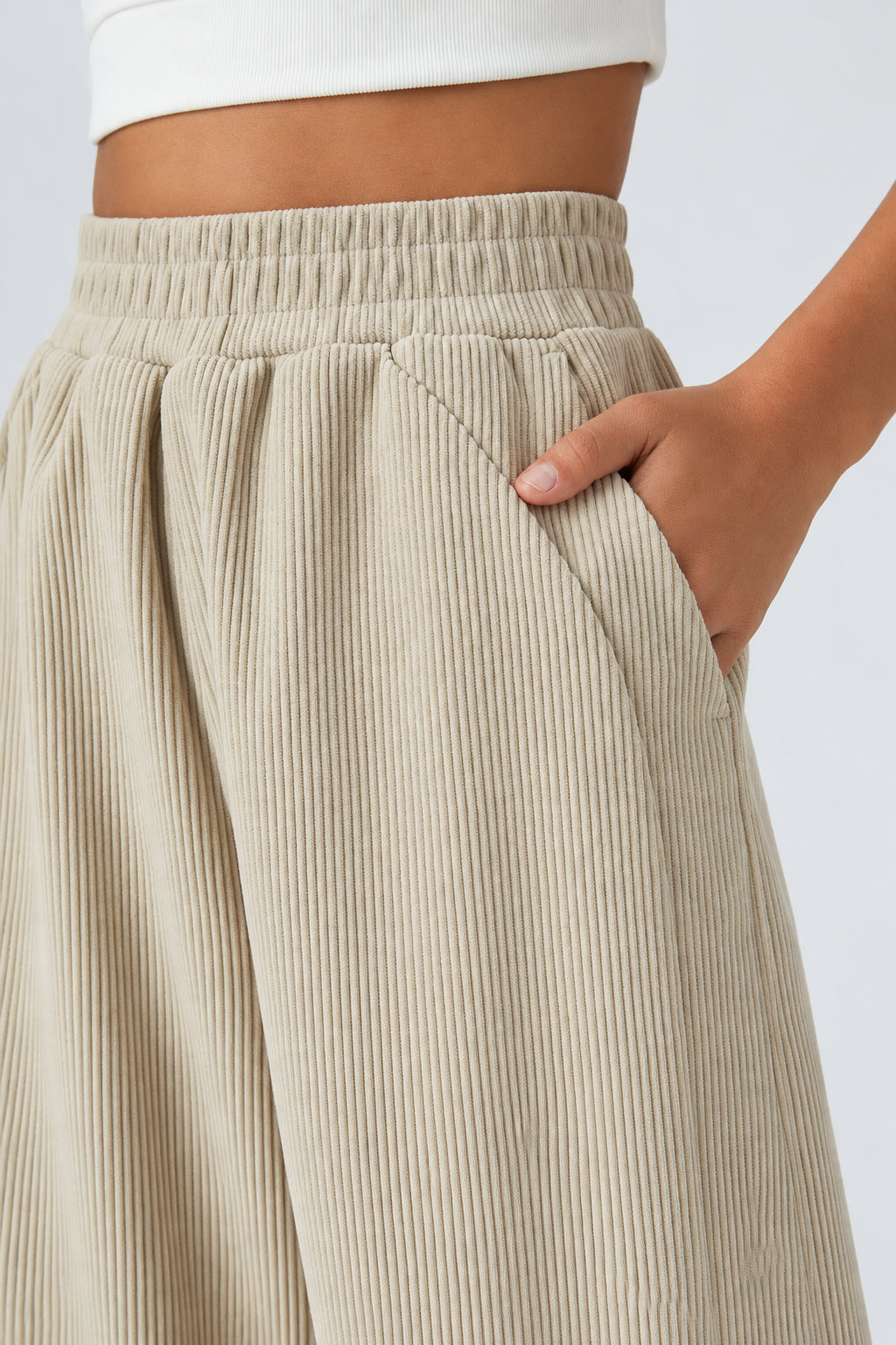 Close-up of a person wearing light beige corduroy wide-leg trousers with an elastic waistband and side pocket, shown against a plain light background. A hand is tucked into the pocket, and part of a white cropped top is visible at the top edge.
