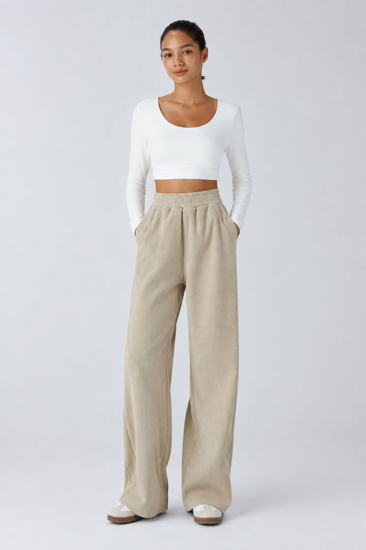 A woman stands against a light gray studio background wearing a white long-sleeve cropped top and beige wide-leg pants. Text in the upper left reads 'Elongates the Legs.