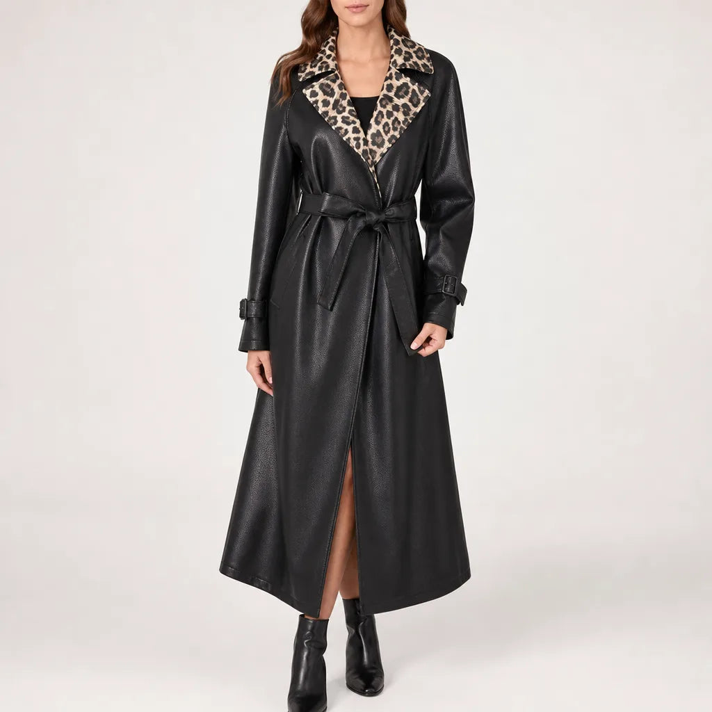 A woman models a long black faux-leather trench coat with a leopard-print collar and matching tie belt, paired with black ankle boots against a plain light gray studio background.