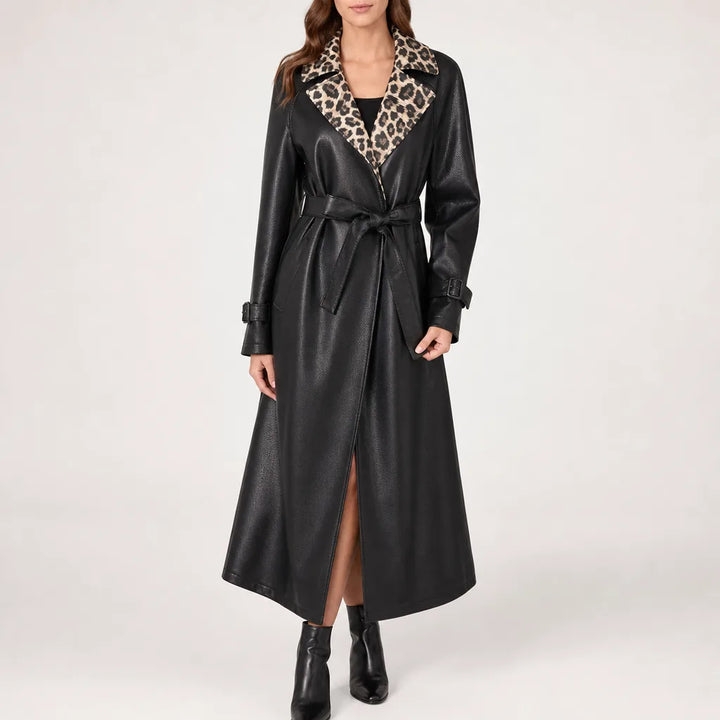 A woman models a long black faux-leather trench coat with a leopard-print collar and matching tie belt, paired with black ankle boots against a plain light gray studio background.
