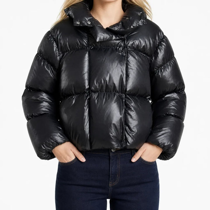 A model wearing a shiny black cropped puffer jacket with a high collar and horizontal quilting, paired with dark blue jeans, against a plain white background.